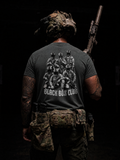A man viewed from behind wearing a dark gray t-shirt with a black and white graphic of three soldiers and the text 'BLACK BOX CLUBS'. He is also wearing tactical gear, including a camouflage helmet, a rifle over his shoulder, and a tactical belt with pouches, set against a very dark background.
