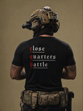 A man viewed from behind wearing a black t-shirt with the slogan 'close quarters battle' printed on the back in white and red, paired with tactical gear and a camouflage utility belt against a solid tan background.