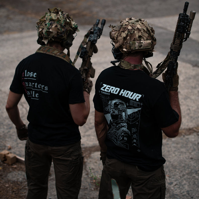 Two people in tactical gear and camouflage helmets seen from behind, standing outdoors on a dirt road and holding rifles. Both are wearing black graphic t-shirts; the left shirt has white text reading 'Iose martyrs' and the right shirt has a large graphic with 'ZERO HOUR' printed across the back.