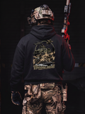 A person shown from behind wearing a black hoodie with a large tactical graphic print on the back, paired with camouflage cargo pants and a tactical helmet, holding a red and black rifle against a dark background.