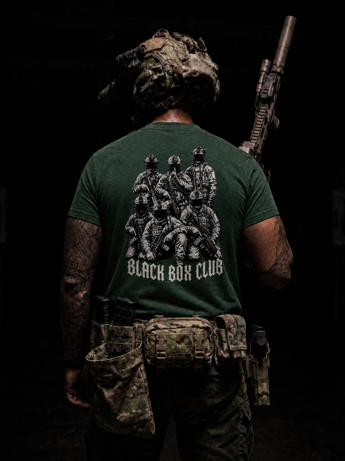 A man viewed from behind wearing a dark green tactical t-shirt with a large black graphic of soldiers and the text 'BLACK BUCK CLUB' on the back, paired with camouflage gear against a dark background.