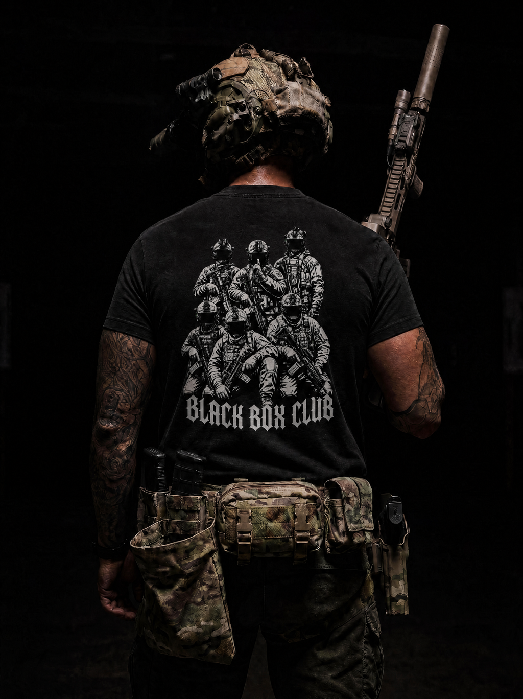 A man viewed from behind wearing a black graphic t-shirt with a white print of soldiers and the text 'BLACK BOX CLUB'. He is also wearing tactical gear, including a camouflage helmet, a rifle over his shoulder, and a tactical belt pouch, set against a very dark background.