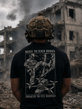 A man viewed from behind wearing a black graphic t-shirt with a white military-themed print and helmet, standing in front of a ruined concrete building with smoke in the background.