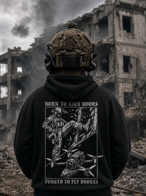 A person viewed from behind wearing a black hoodie with a large white graphic print on the back, standing in front of a war-torn ruined building. The hoodie text reads 'BORN TO KICK DOORS' and 'FORCED TO FLY DRONES.