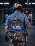 A person at a shooting range seen from behind, wearing a blue graphic T-shirt with a drone and the words 'EXPRESS DELIVERY' on the back, camouflage pants, and a tactical helmet.