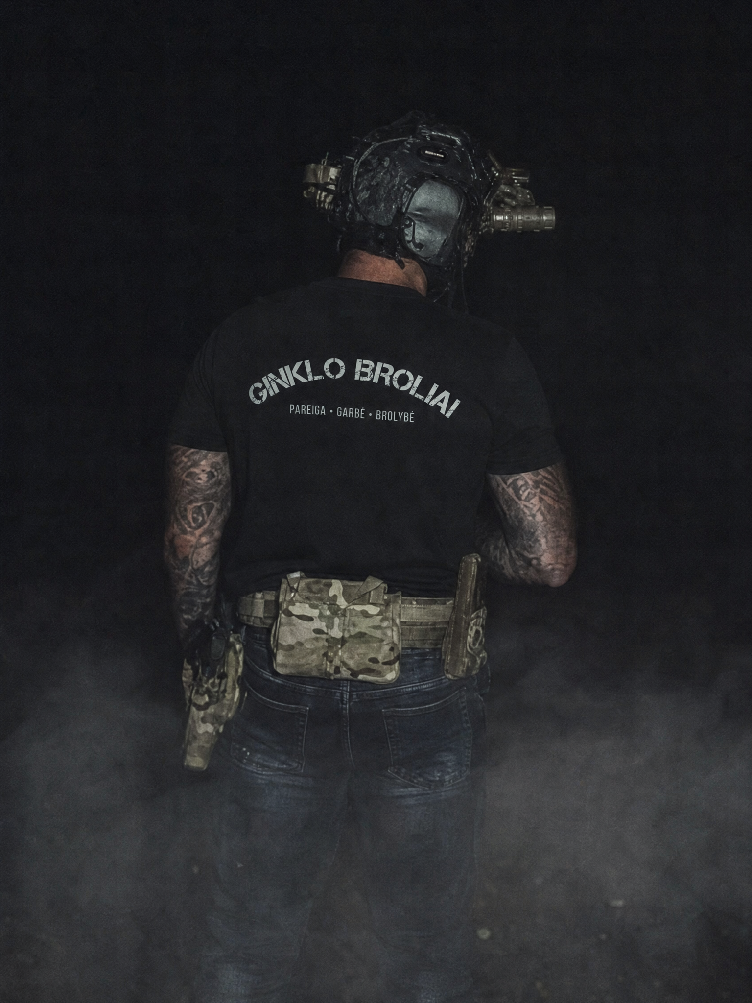 Person wearing a black t-shirt with 'Ginklo Broliai' text, tactical belt, and helmet in a dark setting.