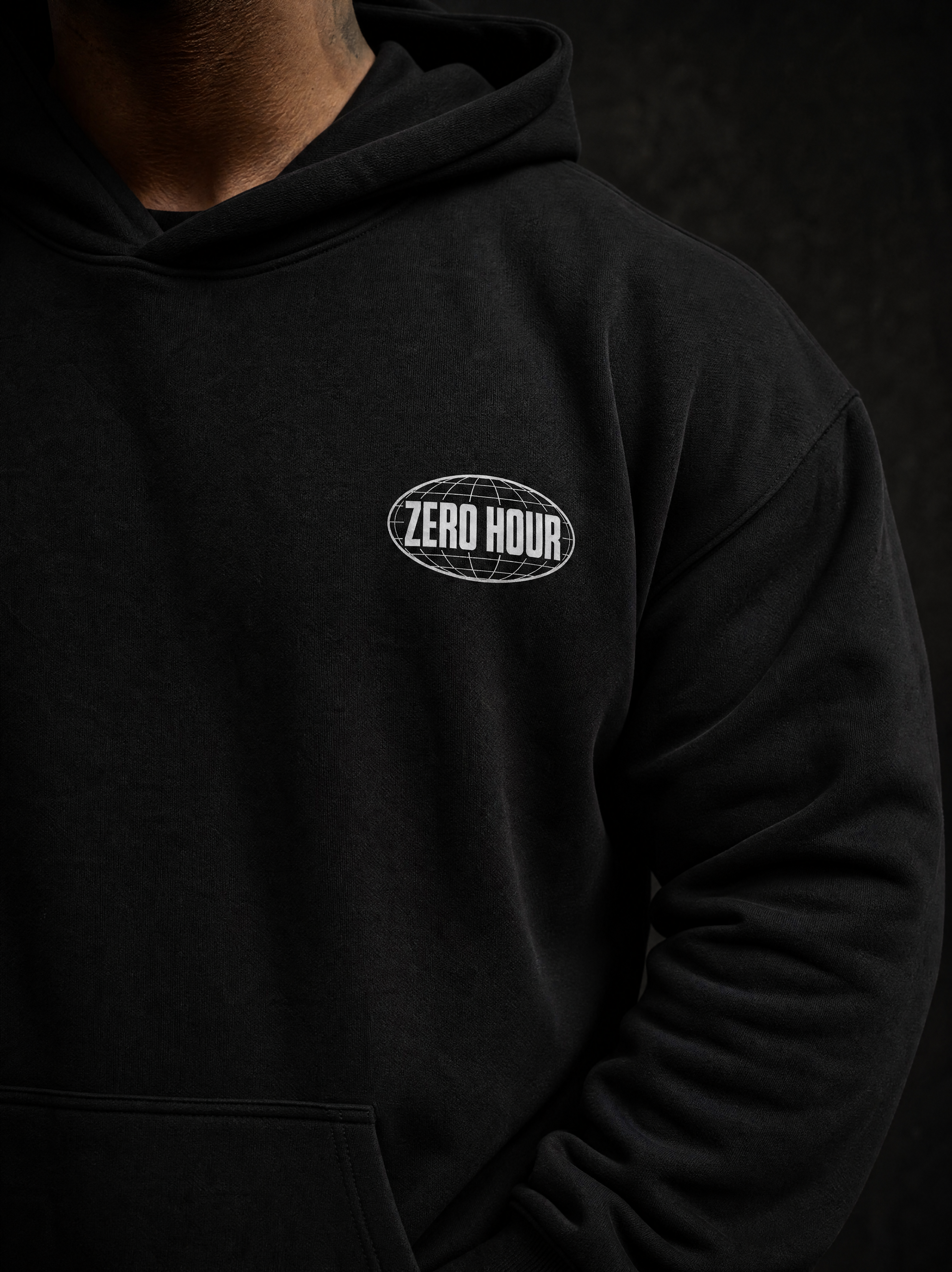A close-up of a person wearing a black pullover hoodie with a white oval 'ZERO HOUR' logo printed on the left chest, set against a dark textured background.