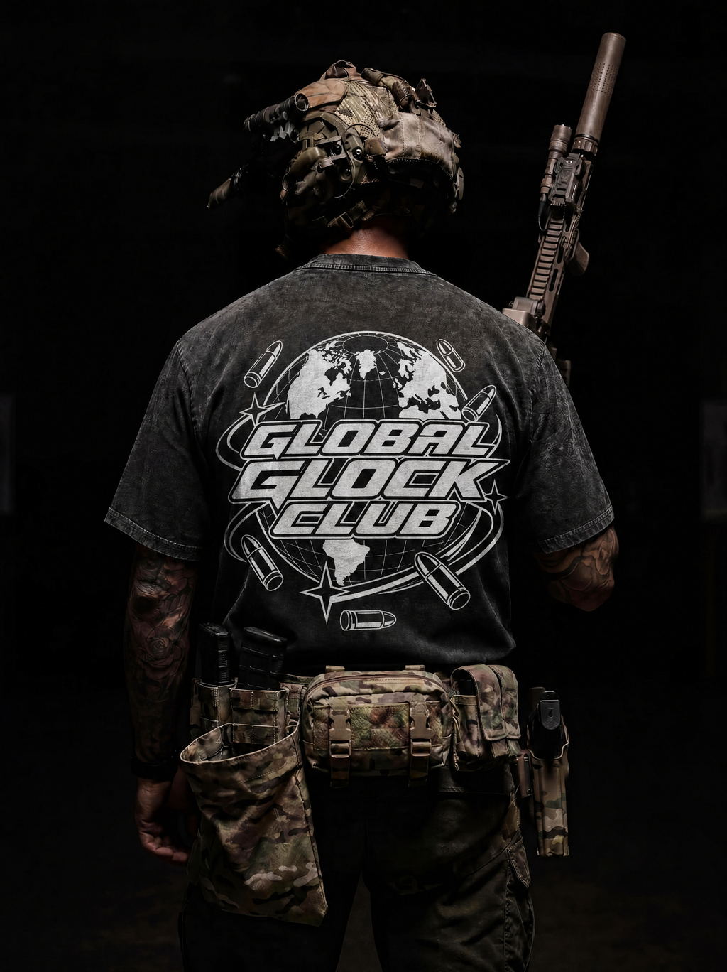 A man viewed from behind wearing a black Global Glock Club graphic t-shirt, tactical gear, and a rifle over his shoulder, set against a dark background.