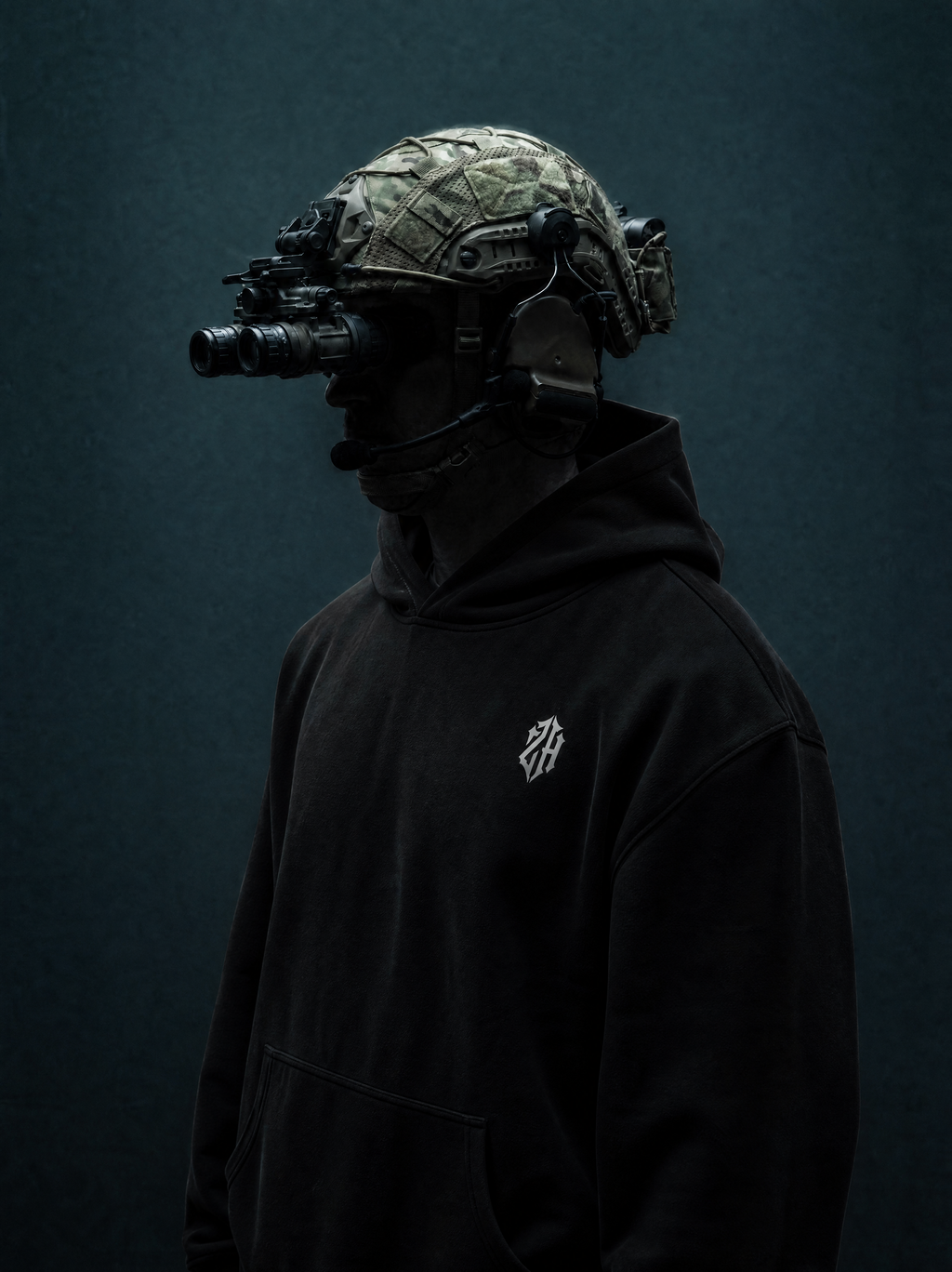 Person wearing a black hoodie with a logo and a camouflage helmet with night vision goggles on a dark background