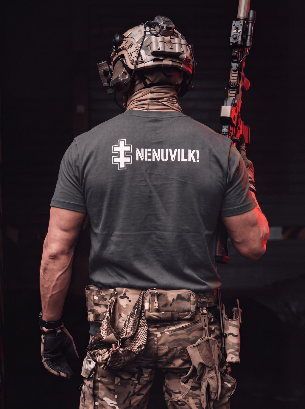 A man viewed from behind wearing a dark gray t-shirt with 'NENUVILK!' and a white cross graphic printed on the back, paired with camouflage tactical pants and a helmet, holding a rifle in a dark setting.