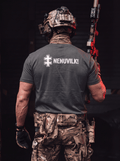 A man viewed from behind wearing a dark gray t-shirt with 'NENUVILK!' and a white cross graphic printed on the back, paired with camouflage tactical pants and a helmet, holding a rifle in a dark setting.