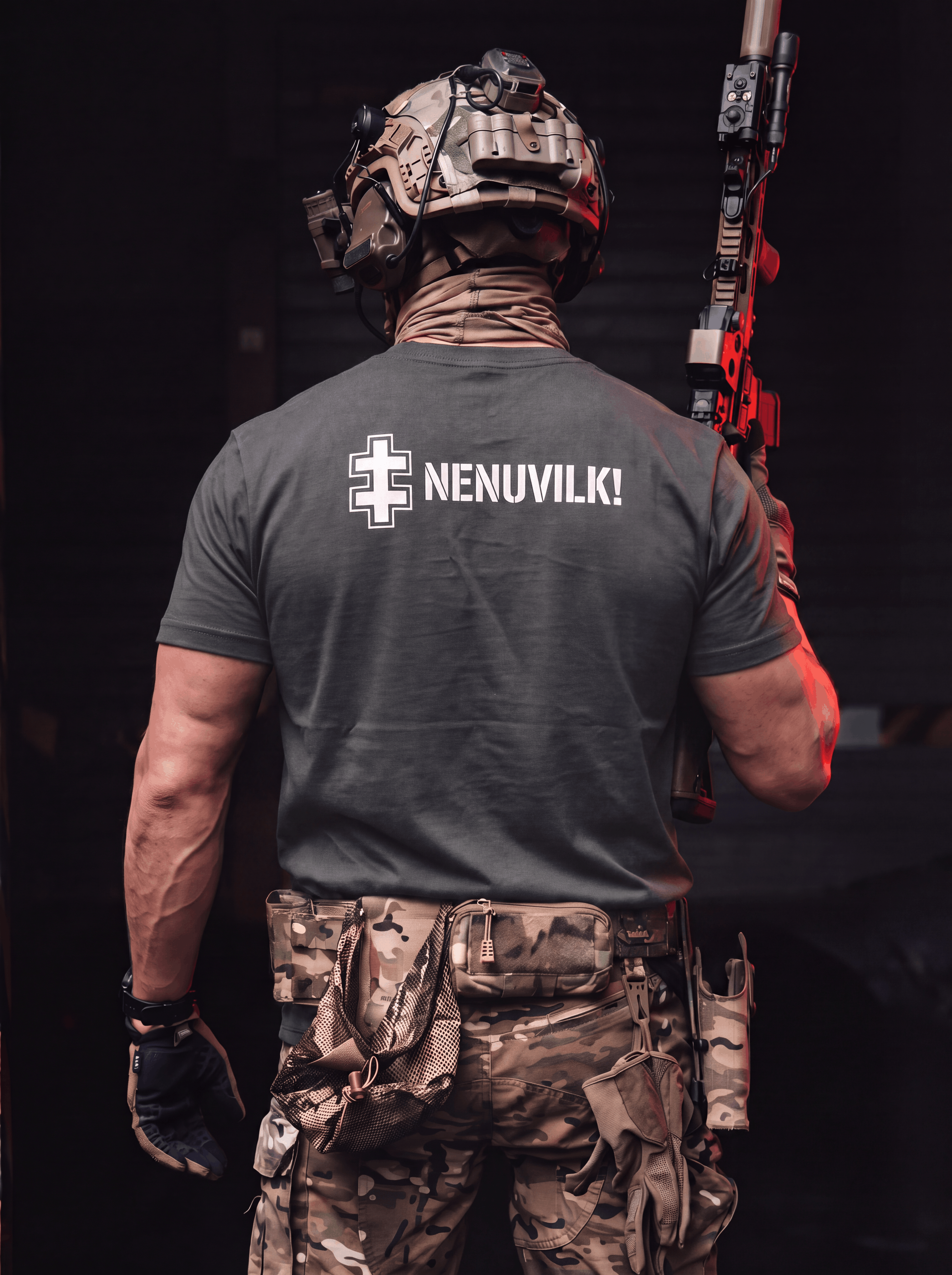 A man viewed from behind wearing a dark gray t-shirt with 'NENUVILK!' and a white cross graphic printed on the back, paired with camouflage tactical pants and a helmet, holding a rifle in a dark setting.