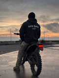 Person wearing a 'zero hour' hoodie standing next to a motorcycle with wind turbines in the background.