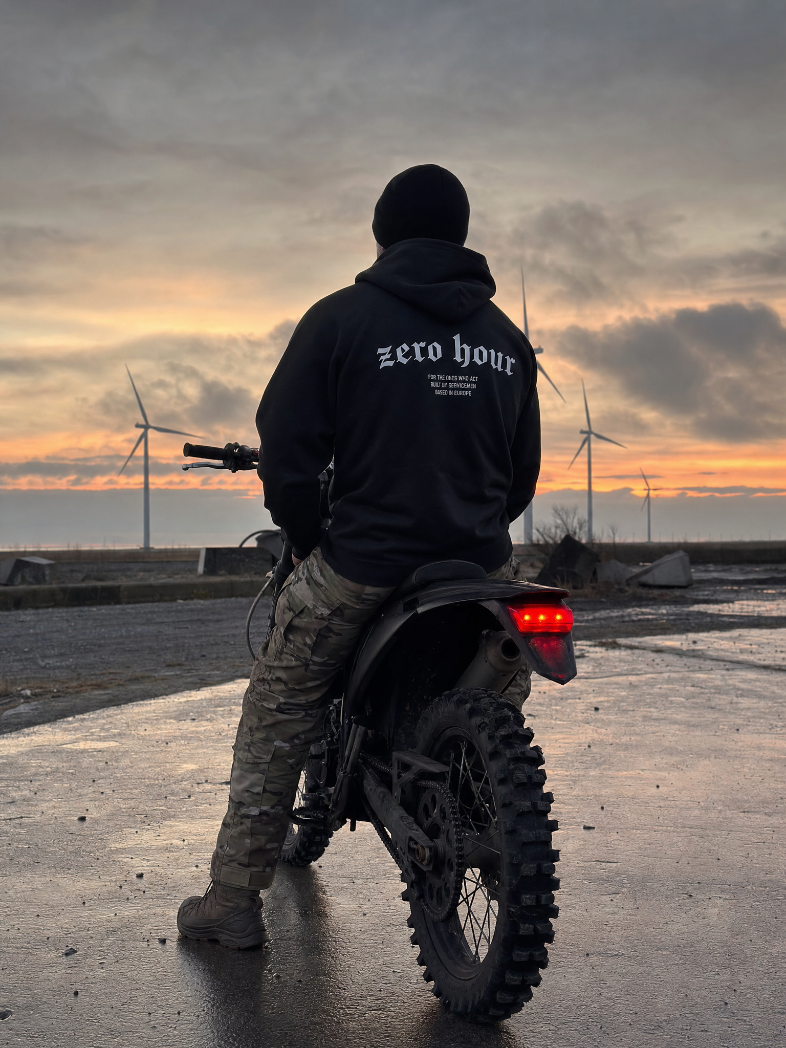 Person wearing a 'zero hour' hoodie standing next to a motorcycle with wind turbines in the background.