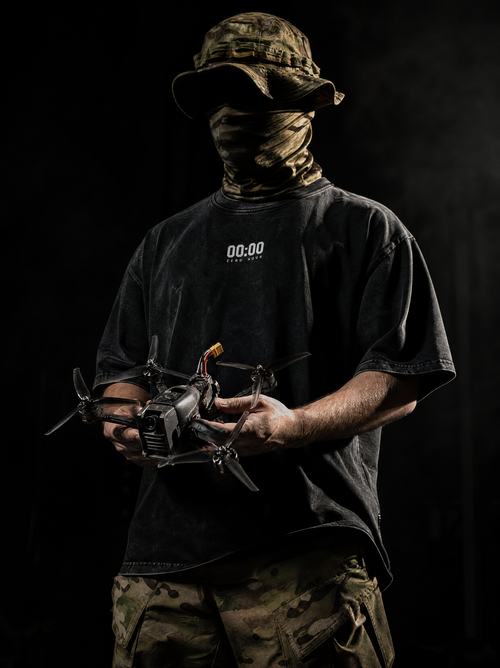 Person wearing a black t-shirt with '00:00' text, holding a drone against a dark background.