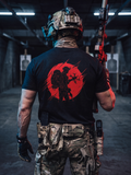 A person viewed from behind wearing a black graphic T-shirt with a large red circular soldier silhouette print, camouflage tactical pants, a helmet, and gloves, standing in a dim indoor shooting range while holding a rifle.