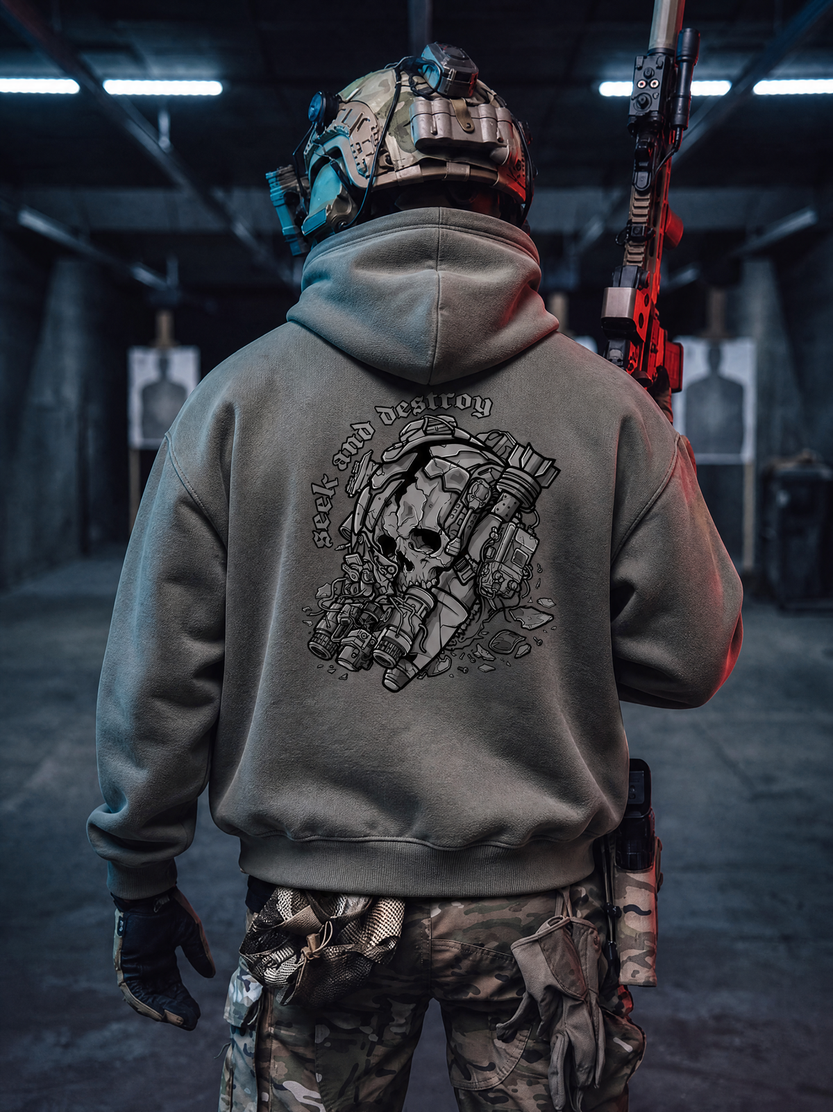 A person viewed from behind wearing a gray hoodie with a large black tactical skull graphic and the words 'Seek and Destroy' on the back, standing in a dim indoor shooting range holding a rifle.