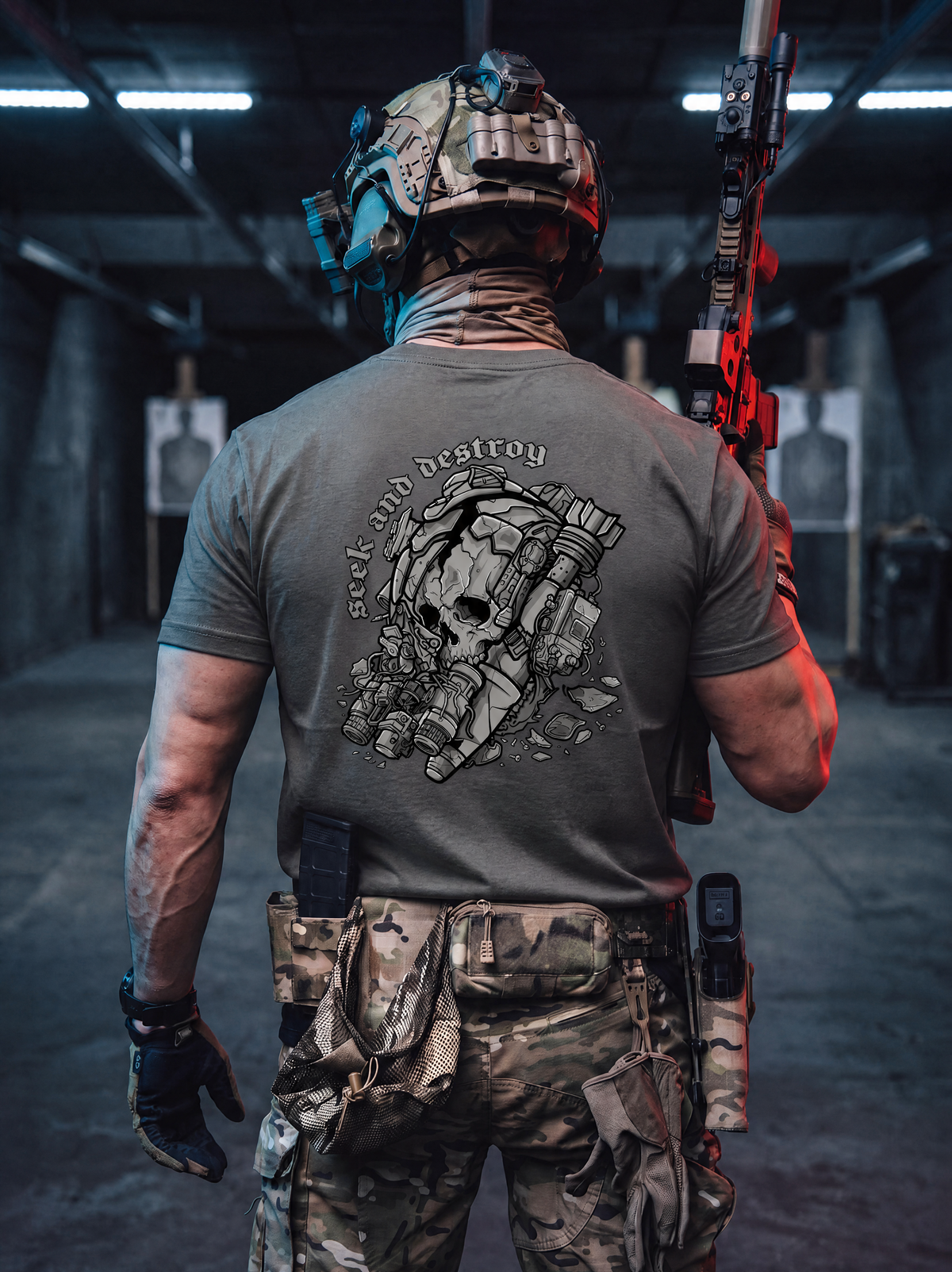 A person standing in a shooting range wearing a gray t-shirt with a large skull and rifle graphic on the back, camouflage pants, and a tactical helmet, holding a rifle.